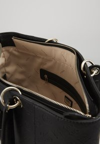 Schwarze Handtasche mit strukturiertem Äußeren, goldfarbenen Beschlägen und einem beige gefütterten Innenraum, der eine Reißverschlusstasche und ein wiederholtes Logo-Muster aufweist.