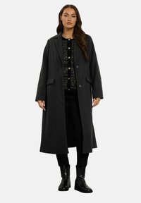 Kobieta w długim ciemnym płaszczu, nosząca czarny sweter zapinany na guziki, czarne spodnie i czarne botki na obcasie, stojąca na jednolitym tle.