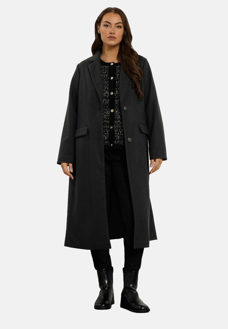 Kobieta w długim ciemnym płaszczu, nosząca czarny sweter zapinany na guziki, czarne spodnie i czarne botki na obcasie, stojąca na jednolitym tle.