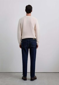 Suéter de punto crema, jeans de mezclilla azul, zapatos marrones, vista desde la parte de atrás. El suéter tiene puños acanalados y un diseño minimalista.