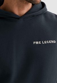 Hombre con sudadera con capucha azul marino oscuro con "PME LEGEND" bordado en el pecho, primer plano que muestra la barba y el cuello.