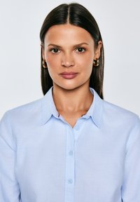 Sieviete ar taisniem tumšiem mati, valkājot zelta riņķa auskarus un gaiši zilu pogas aizdares kreklu, tieši skatās kamerā uz vienkārša fona.