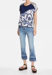 Top blu navy e bianco con accenti argentati sulle maniche, abbinato a jeans ricamati azzurri e sandali neri con dettagli rossi.