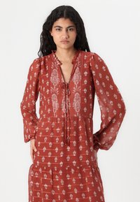 Robe à motifs rouges avec un décolleté à nouer, un col froncé et des manches longues. Elle présente des motifs floraux blancs et une jupe à étages.