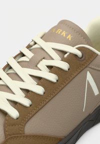 Világosbarna sneaker sima felsőrésszel, krémszínű fűzőkkel és sötét gumitalppal. Jellemzője egy arany logó és egy geometrikus fehér akcentus.