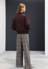 Giacca bordeaux con zip e collo, abbinata a pantaloni a gamba larga a quadri in tonalità di grigio e crema, in piedi su uno sfondo monocromatico.