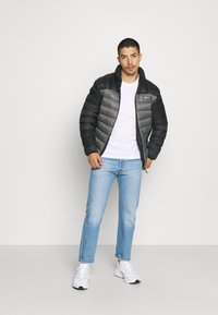 Giubbotto imbottito in nero e grigio con cuciture orizzontali, chiusura con zip e logo sul petto, abbinato a jeans azzurri chiari e sneakers bianche.