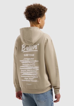 Tienerjongen met krullend haar, gekleed in een beige hoodie met witte tekst "Ballin Surf Tour" en doorgestreepte surfbestemmingen op de achterkant, zijwaarts kijkend.