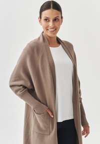 Cardigan beige ouvert, tissu tricoté texturé, manches longues avec des poignets côtelés, deux poches avant, porté sur un haut blanc, silhouette ajustée.