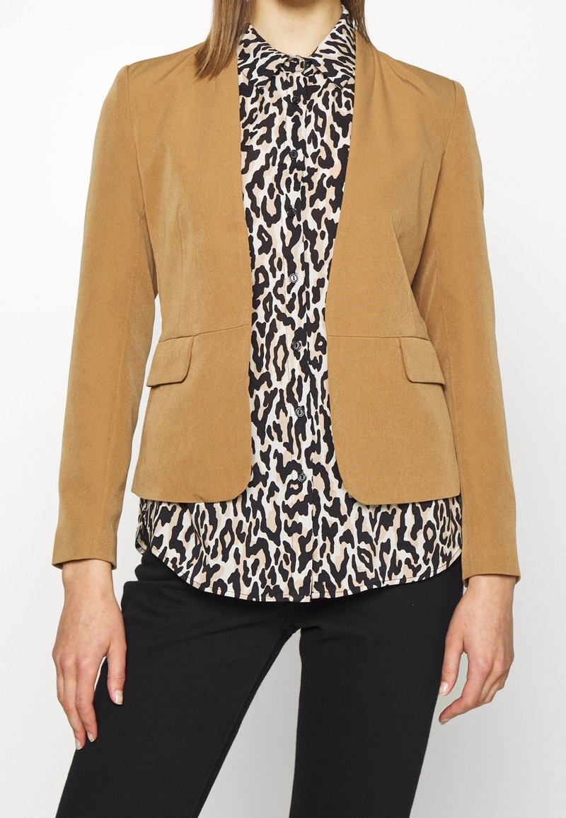 Femme portant un blazer couleur beige sur une chemise boutonnée imprimée léopard noir et blanc avec un pantalon noir, debout devant un fond uni.