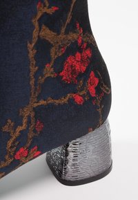Chaussure en tissu foncé avec un motif floral rouge et marron, dotée d’un talon bloc métallique texturé.
