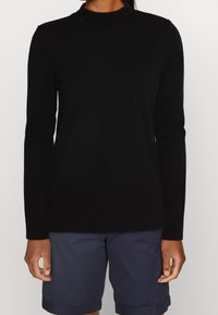 Schwarzes langärmeliges Turtleneck-Oberteil aus weichem Stoff mit taillierter Passform und dezent geribbeltem Halsausschnitt, kombiniert mit marineblauen Shorts.