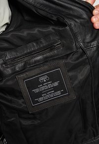 Intérieur d'une veste en cuir noire, présentant une poche zippée et une étiquette en tissu avec des instructions d'entretien. Texture lisse et design épuré.