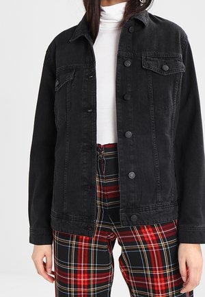 Femme portant une veste en jean noire boutonnée, un col roulé blanc et un pantalon à carreaux rouge taille haute.