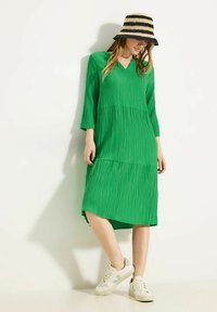 Grünes, strukturiertes Midi-Kleid mit V-Ausschnitt und dreiviertel langen Ärmeln. Kombiniert mit weißen Sneakers mit grünen Akzenten.