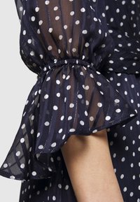 Blouse bleu marine transparente avec pois blancs, manches élastiquées à volants, et rayures subtiles, mettant en valeur une texture légère et aérée.