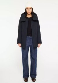 Zwarte gewatteerde jas met capuchon, vooraan met een rits, zijzakken, gecombineerd met een losse blauwe denim broek en een witte gebreide beanie.