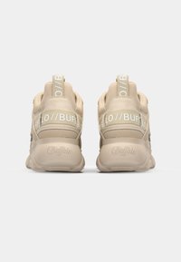 Beige sneakers with textured fabric and smooth accents, featuring a pronounced sole and branded heel tab in white lettering.