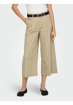 Pantalon beige large court avec plis et poches latérales, associé à une ceinture noire et des chaussures plates tissées noires.