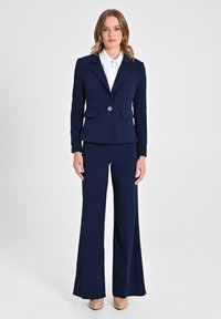 Costume deux pièces bleu marine comprenant un blazer ajusté avec un bouton doré unique et un pantalon évasé, complété par une chemise à col blanc.