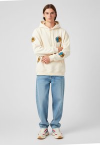 Sudadera crema con motivos de sol en amarillo y azul; combinada con jeans azul claro y zapatillas blancas con detalles en rojo y azul.