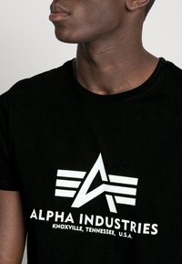 Black cotton t-shirt featuring a large white graphic with the text "ALPHA INDUSTRIES" and a stylized logo centered on the front.
