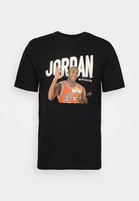 Fekete pamut póló, amelyen egy narancssárga mezben játszó kosárlabdázó grafikus nyomata található, erőteljes fehér szöveggel: "JORDAN."