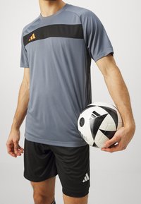Šedé sportovní tričko s černým vodorovným pruhem a oranžovým brandingem, spárované s černými šortkami, držící černobílý fotbal.