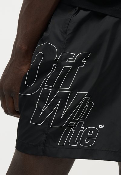 Άτομο σκουρόχρωμο που φορά μαύρα σορτς με μεγάλο λευκό περίγραμμα "Off-White" στο πλάι κοντά στο μηρό.