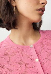 Rosa gestrickte Strickjacke mit einem Blattmuster, rundem Ausschnitt und Knopfverschluss mit einem hellen Knopf. Strukturierter Stoff.