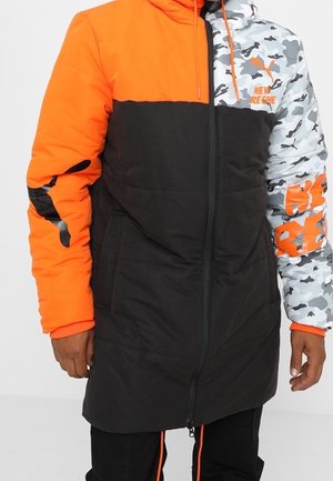 Vadderad jacka med en blandning av svarta, orange och grå kamouflagemönster, med dragkedja fram och logodetaljer på ärmen.