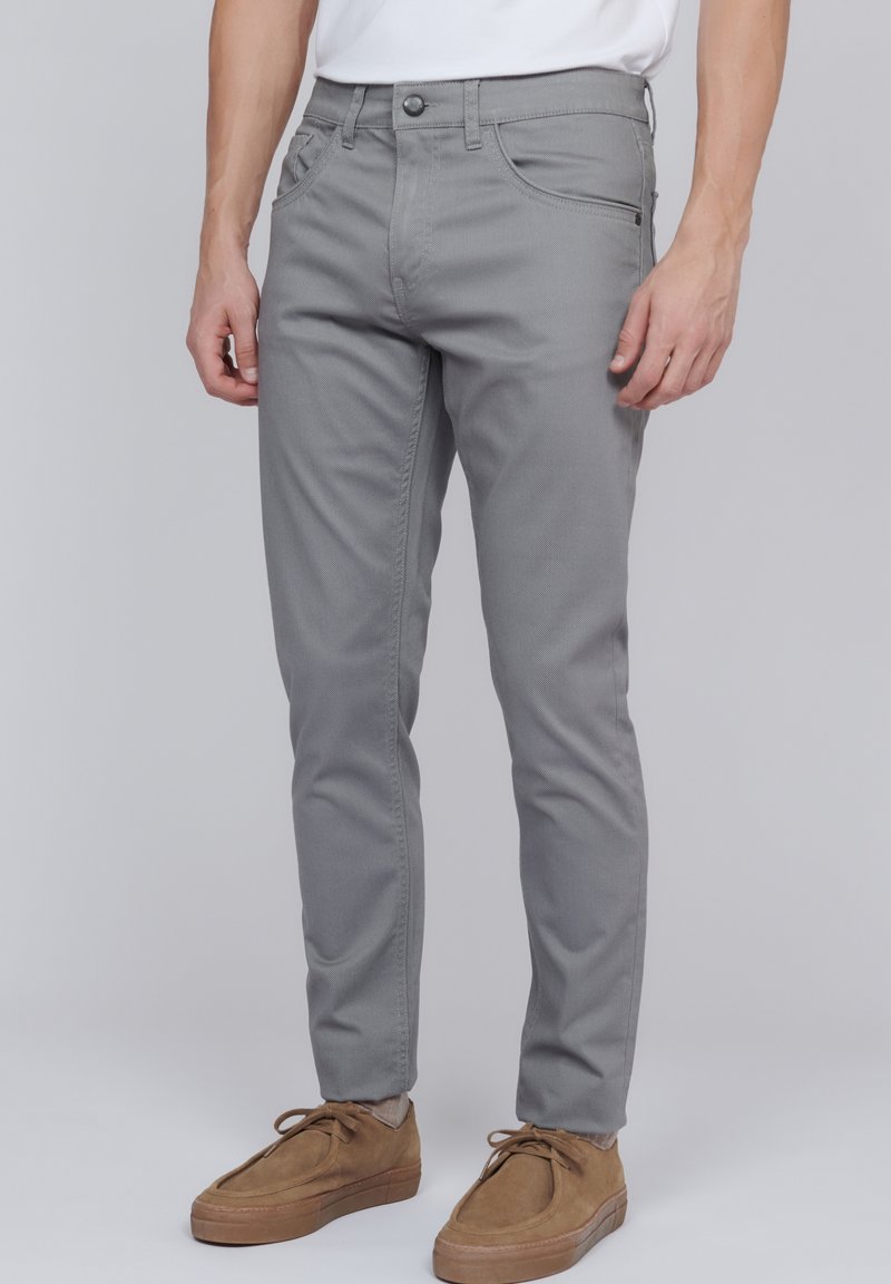 Pantalon gris à coupe slim en tissu tissé, avec poches avant et fermeture à bouton. Associé à des chaussures en daim marron.