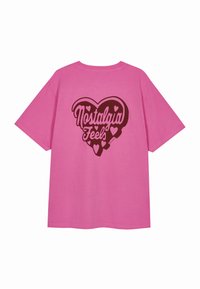 Rozā kokvilnas t-krekls ar īsām piedurknēm, kas izceļas ar lielu sirds grafiku. Teksts "Nostalgia Feels" ir veidots tumši brūnā krāsā un to ierobežo mazākas sirdis.
