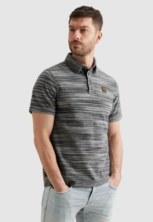 Homme portant un polo gris rayé à manches courtes et un jean bleu clair, debout les mains dans les poches, regardant légèrement sur le côté.