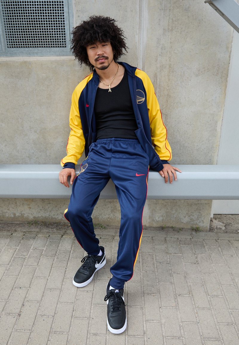 Modrý a žltý športový komplet Nike s červenými akcentami, čierny tielko, čierne tenisky a reťazový náhrdelník, sediaci na lavičke.