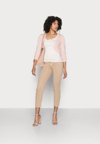 Light pink cardigan over a white top, beige fitted pants, and nude high-heeled sandals. Model holding a matching handbag. Plain background.