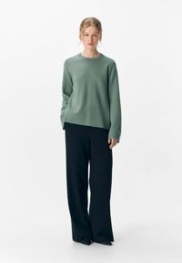 Jeune femme se tenant debout, portant un pull à manches longues vert sauge et un pantalon large noir, devant un fond blanc uni.