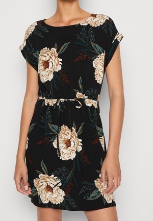 Kvinde iført kort sort kjole med stort beige blomsterprint og bundet taljebælte, stående mod en ensfarvet lys baggrund.