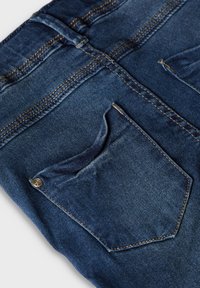 Dark blue denim jeans feature a back pocket with a subtle texture, contrasting stitching, and a metal rivet accent on the pocket.