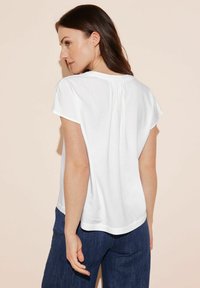 Top bianco a maniche corte con una vestibilità rilassata, caratterizzato da un dettaglio arricciato nella parte posteriore del collo e un orlo curvo. Abbinato a jeans blu scuro.