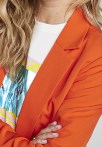 Blazer orange avec un revers structuré, associé à une chemise blanche ornée d'un motif graphique coloré et métallique. Tissu doux à la texture lisse.