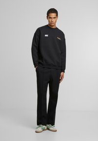 Sudadera negra con logos impresos, combinada con pantalones negro ajustados y zapatillas blancas con acentos verdes. Presenta cuello redondo y corte relajado.