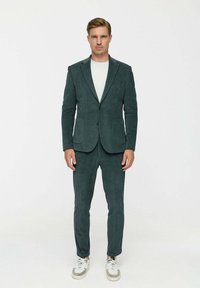 Zöld kordbársony öltöny, illeszkedő blézerrel és távozó nadrággal, világos pólóval és fehér tornacipővel párosítva. Jellemzője a texturált függőleges csíkok.