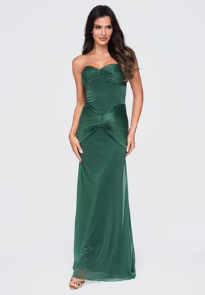 Femme aux longs cheveux foncés portant une robe de soirée verte foncée pailletée sans bretelles et des talons ouverts dorés, debout devant un fond clair uni.