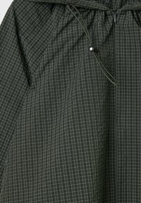 Tissu vert foncé avec un motif à carreaux blancs, doté d'une taille élastique froncée et d'une fermeture éclair noire avec cordon et curseur.