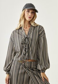 Blusa a rayas, corta en negro, beige y blanco, con un delantero con cordones, mangas abullonadas y tela suave. Combinada con una falda a juego.