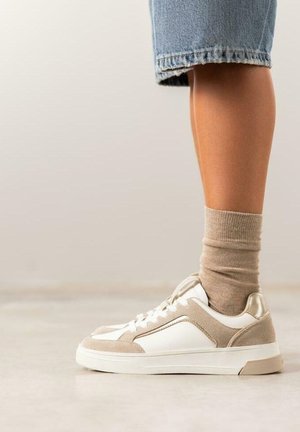 Zapatillas blancas y beige con parte superior de ante y piel, suela blanca plana y detalles reflectantes. Combinadas con un calcetín beige y shorts de mezclilla.