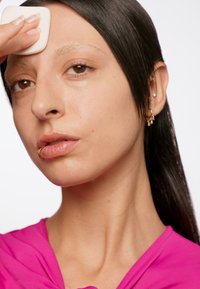 Gros plan sur un visage à la peau marron moyen, portant un haut rose. Une main tient une éponge blanche contre le front, mettant en valeur la texture de la peau naturelle.