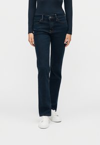 Άτομο που φοράει σκουρο μπλε τζιν παντελόνι με ίσια γραμμή, μπλε σκούρο μπλουζάκι με μακριά μανίκια και λευκά πλατφόρμα αθλητικά παπούτσια, στέκεται μπροστά σε απλό φόντο.