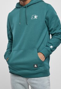Sudadera con capucha color verde azulado hecha de una mezcla suave de algodón, con un bolsillo frontal y capucha con cordón ajustable. Logo impreso en el pecho y la manga.